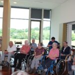 Več starejših ljudi sedi na stolih in invalidskih vozičkih.