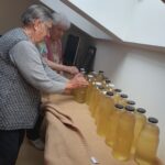 Dve starejši ženski stojita zraven mize na kateri so steklenice bezgovega soka.