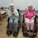 Dve starejši ženski na invalidskih vozičkih. Leva v roki drži mikrofon.
