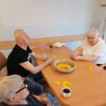 Starejši moški in dve ženski sedijo pri mizi in delajo rumene papirnate kroglice.