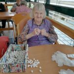 Starejša ženska sedi pri mizi in dela bele papirnate kroglice.