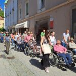 Več ljudi na invalidskih vozičkih s spremljevalci se sprehaja po ulici v mestu.