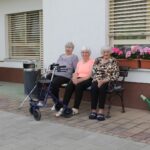 Tri starejše ženske sedijo na klopci pred stavbo.