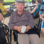 Starejša gospa s klobukom sedi na stolu in v rokah drži lonček.