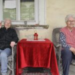 Dve starejši gospe sedita na lesenih stolih, v sredi pa je mizica z rdečim prtom.