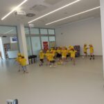 Skupina otrok v rumenih majicah in kapah s šiltom pleše v paru.
