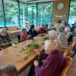 Več starejših ljudi sedi za mizo in pripravlja zelišča.
