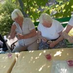 Štiri starejše ženske sedijo za mizo na prostem, obdane z zelenjem. Vsaka od njih ima v rokah skodelico in žličko, uživajo v ledeni kavi ali sladoledu. Na mizi so tudi kozarci z rožnato pijačo.