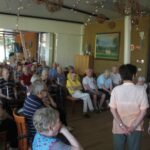 Velika skupina starejših ljudi sedi v prostorni sobi in pozorno spremlja nastop. Soba je okrašena s slikami, visečimi okraski in ima velika okna.