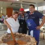 Trije ljudje, med njimi ženska v beli majici, moški v rdeči kapi in moški v modrem predpasniku, stojijo ob velikem kotlu s pečenimi kosi mesa. Ženska meša vsebino kotla, moška pa se nasmejana objemata.