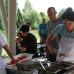 Skupina ljudi, ki se vrstijo ob mizi s postrežbo hrane na samopostrežnem bifeju. Ženska v svetlomodrem predpasniku si naloži hrano na krožnik. Na voljo so različne jedi, vključno z narezanim paradižnikom.