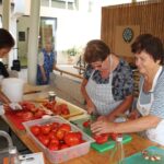 Tri ženske, oblečene v predpasnike, režejo paradižnike na mizi. Na mizi so posode s paradižniki, nekatere že narezane na koščke. V ozadju je več ljudi, ki sodelujejo pri pripravi hrane.