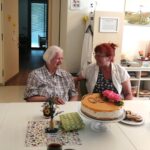 Dve ženski sedita ob mizi na kateri je torta z napisom "90".