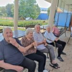Trije starejši moški in ženska sedijo na klopci na prostem in jedo sladoled.