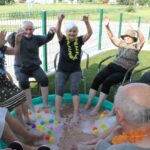 Več starejših ljudi sedi v krogu in namaka noge v bazen, pri tem imajo dvignjene roke.