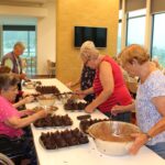 Več starejših žensk je ob mizi na kateri pripravljajo muffine.