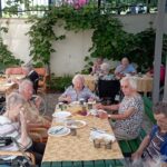 Več starejših ljudi sedi za mizo na prostem in je zajtrk.