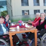 Štiri starejše ženske sedijo za mizo na prostem na kateri je pripravljen zajtrk.