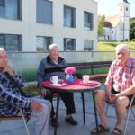 Trije starejši moški sedijo za mizico na prostem na kateri so skodelice.
