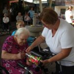 Starejša gospa v roza obleki z vzorcem cvetlic izžreba listek iz steklenega kozarca, ki ji ga ponuja ženska v beli majici. Dogajanje poteka na terasi, v ozadju pa so drugi starejši ljudje, ki sedijo in se pogovarjajo.