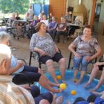 Skupina starejših se druži na terasi, kjer si namakajo noge v bazenu z vodo in rumenimi žogicami. V ozadju se drugi stanovalci družijo ob mizah in uživajo v prijetnem poletnem dopoldnevu.