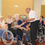 Starejši moški stoji pred sedečo starejšo žensko in ji izroča šopek rož, medtem ko jo drži za roko. Okoli njiju je skupina starejših oseb, ki spremljajo dogodek z nasmeški na obrazih, nekateri so videti ganjeni.