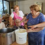 Skupina starejših žensk med izdelavo ognjičevega mazila. Ena od žensk meša sestavine v loncu, medtem ko druga nežno dodaja dodatne sestavine. Dogajanje spremlja negovalka v roza halji.