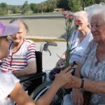 Otrok v svetli majici in roza kapi izroča rožnati nagelj starejši ženski, ki sedi v invalidskem vozičku. Oba se nasmehneta, v ozadju pa sta še dve starejši ženski, ki ju spremljata.