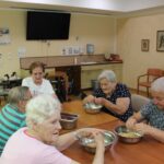 Skupina starejših žensk sedi za mizo in aktivno pripravlja sladico. Vsaka ima pred seboj kovinsko posodo s sestavinami. Na sliki je prikazan domač, prijeten prostor z lesenim pohištvom in televizijo na steni.