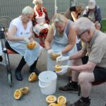 Starejši moški in ženske sedijo na stolih na prostem, oblečeni v plastične predpasnike in rokavice, medtem ko čistijo buče. Nekateri držijo polovico buče in s pomočjo žlic odstranjujejo semena.