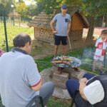 Skupina starejših ljudi sedi zunaj na vrtu, medtem ko pečejo koruzo na žaru. V bližini je tudi mlajši moški in deček, ki opazujeta dogajanje.