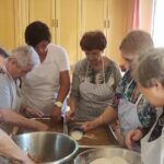 Na sliki ženske zbrano pripravljajo sestavine za sladico, natančno odmerjajo sestavine in jih skupaj urejajo v posodo.