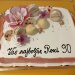 Na torti bele barve z roza robovi je napis "Vse najboljše Rozi 90". Na vrhu torte so okraski iz makronov, sladkornih rož in kroglic. Tortni pladenj je postavljen na rumeno mizo.