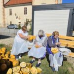 Tri starejše ženske sedijo na klopi ob velikih kupih buč, vsaka ima v rokah bučo, ki jo treblji. Na klopi imajo posode za shranjevanje bučnih semen, dogodek poteka na prostem pred stavbo.