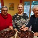 Tri starejše gospe stojijo pred dvema skledama, polnima kuhanih kostanjev. Gospe so nasmejane in sodelujejo v dejavnosti z okusnimi kostanji.