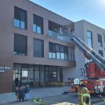 Na parkirišču pred veliko moderno stavbo so gasilski tovornjaki. Na strehi enega tovornjaka je raztegnjena lestev, po kateri se gasilci vzpenjajo na balkon. Gasilci v uniformah so aktivni, pripravljajo opremo, na tleh je razvita cev za gašenje požara. Nekaj ljudi, vključno z otroki, opazuje dogajanje.