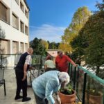 Skupina starejših ljudi skupaj s prostovoljci sadi rastline v lonce na prostorni terasi. V ozadju je stavba z zelenimi drevesi, ki jih obsije sonce, kar ustvarja prijetno jesensko vzdušje.