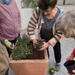 Starejša ženska z rjavimi lasmi sadi cvetlice v velik lončen cvetlični lonec, obdana z drugimi starejšimi udeleženkami, ki opazujejo. Okoliška vegetacija prispeva k prijetnemu in umirjenemu vzdušju na prostem.