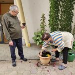 Moški in ženska skupaj sadita cvetlice v majhen okrogel lonček. Moški drži prazen črn plastični lonček, medtem ko ženska posadi cvetlice. Oba sodelujeta v sajenju, obkrožena z zelenimi rastlinami in vegetacijo.