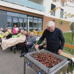 Na dvorišču pred stavbo moški stoji pred velikim žarom, na katerem peče kostanj. V ozadju več starejših ljudi sedi na ležalnikih, zaviti v odeje, in uživajo v družabnem dogodku. Vzdušje je jesensko in toplo.