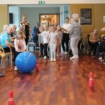 Mlajša deklica z modro žogo igra bowling v zaprtem prostoru, kjer jo spremlja skupina starejših oseb in nekaj otrok. Starejši sedijo na stolih ali v invalidskih vozičkih ob straneh, medtem ko deklica usmerja žogo proti rdečim kegljičem na koncu proge. V ozadju stojijo skrbniki in spremljajo dogajanje.