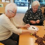 Dve starejši ženski sedita za mizo, medtem ko pripravljata kostanje. Ena jih lušči s pomočjo noža, druga pa jih pripravlja na krožniku. Oba krožnika imata rdeč obod.