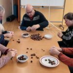 Skupina starejših moških sedi za veliko mizo in lupijo kostanje. Kostanj je raztresen po mizi, nekateri imajo krožnike s kostanji pred seboj. Razporeditev mize je pravokotna in široka.