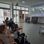 Skupina starejših ljudi sedi v vrstah pred velikim projekcijskim zaslonom, na katerem se predvaja film z napisi. Nekateri gledalci sedijo v invalidskih vozičkih in uživajo v pokovki med gledanjem filma. Na desni strani so mize s prigrizki in pijačo, ki so pripravljeni za postrežbo.