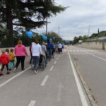 Pogled od zadaj na udeležence sprehoda, ki se premikajo po široki cesti. Mnogi potiskajo invalidske vozičke, nekateri ljudje pa držijo modre balone. Na levi strani so otroci in odrasli, nekateri oblečeni v pisana oblačila, ki se pridružujejo procesiji, medtem ko na desni strani cesto obdajajo drevesa in visoka ograja.