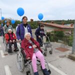 Skupina starejših žensk na invalidskih vozičkih, ki jih vodijo spremljevalke, sodeluje v sprehodu. Nekatere držijo modre balone. Ženske so videti sproščene, spremljevalke pa nasmejane. Sprehajališče poteka ob cesti, v ozadju pa so hiše in zelenje.