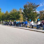 Skupina ljudi, večinoma starejših oseb v invalidskih vozičkih, stoji na robu pločnika. Nekateri imajo v rokah modre balone, sprehod poteka ob cesti, ozračje je sončno in nebo jasno.