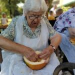 Starejša gospa z belimi lasmi in očali, oblečena v cvetlično bluzo, čisti bučo. Njeno delo je natančno, medtem ko se osredotoča na notranjost buče, ki jo drži v naročju. V ozadju so še drugi udeleženci dogodka na prostem.