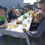 Dolga miza postavljena zunaj, okoli katere starejši moški in ženske uživajo v skupnem obroku. Na mizi so kozarci soka in krožniki hrane, ženska v roza predpasniku pa streže pijačo. Skupina sedi na soncu poleg bele stavbe.