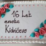 orta z belim premazom in okrasnimi rdečimi vrtnicami ter napisom "16 let enote Kidričevo" v čokoladni glazuri. Elegantno oblikovana torta z nežnimi barvnimi poudarki.